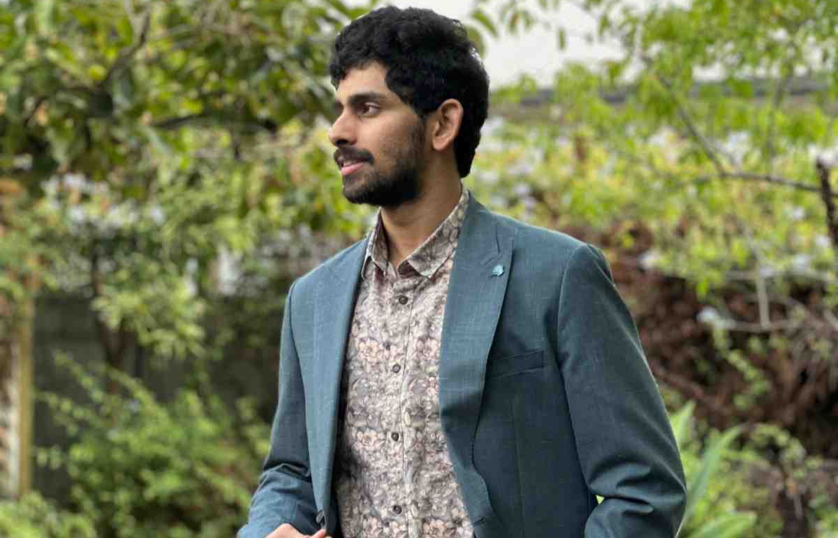 A man with dark hair and a beard is standing outdoors, looking to his left. He is wearing a teal blazer over a floral button-up shirt. The background is filled with green plants giving a natural, garden-like setting.